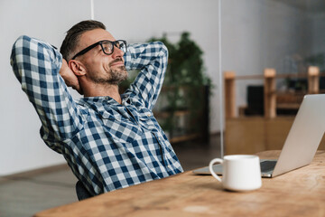 Wall Mural - Pleased unshaven man in eyeglasses resting while working with laptop