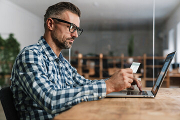 Wall Mural - Focused unshaven man using credit card while working with laptop