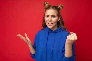 Photo of emotional agressive young beautiful blonde woman with sincere emotions wearing blue pullover isolated over red background with free space and showing fist