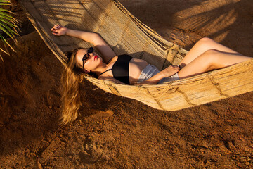 Wall Mural - Young beautiful girl in sunglasses posing while lying on a hammock, beautiful woman resting on vacation at the beach, relax, outdoor portrait