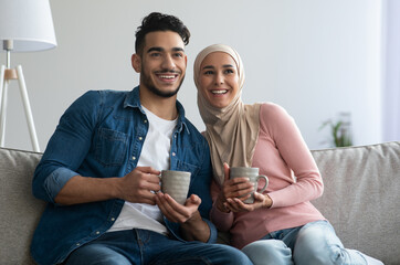 Wall Mural - Happy middle-eastern couple sitting on couch at home, drinking tea