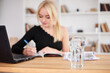 © Natalia - Young blond woman, wearing black top, sitting by office table with huge glass of water on it.Manager at workplace. Work process in company. Healthy drink consumption concept.Water balance.