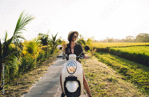 Millennial female travel blogger testing vintage motor transport during summer vacations holding cellphone device for networking, charming hipster girl with smartphone gadget resting on scooter