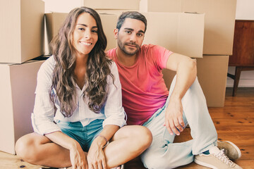 Happy Latin couple in love enjoying moving into new apartment, sitting on floor near heap of carton boxes, looking at camera and smiling. New home or relocation concept