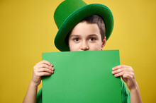 Handsome Boy Dressed In A Leprechaun With A Green Irish Cap Covers His Face With A Sheet Of Green Paper With Copy Space. Saint Patrick's Day Concept.