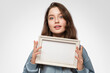 © Snizhana - Young woman with an empty picture frame horizontally in her hand on a light blue background. The frame has a space for your text or photo. Light blue hoodie, blue eyes long hair.
