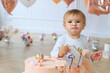 Close up Cute blond baby girl in peach tutu sitting next to on festive background by smashed decorated fondant iced cake with dirty sticky hands and mouth from messy crumb cake. cake smash concept