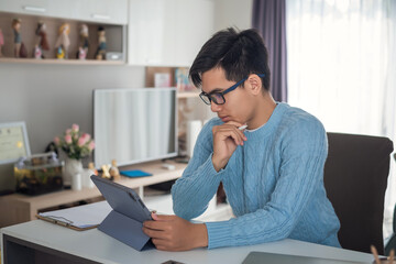 Young Asian man wearing glasses is working on a tablet at home.