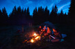 © anatoliy_gleb - Group of four tourists sitting together near campfire, enjoying fresh air near tent with tourists equipment nearby, dark relaxing atmosphere. Evening camping near fire, spruce forest on background.