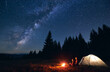© anatoliy_gleb - Young couple hikers sitting near bright burning bonfire and illuminated tourist tent, enjoying camping night together under dark sky full of shiny stars and bright Milky Way, warm summer evening.
