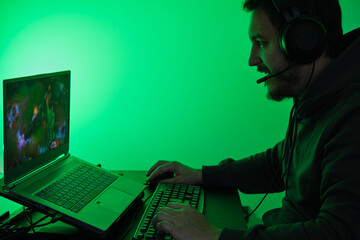 Wall Mural - Gamer wearing headphones, playing video games in his laptop. Green light style.