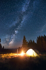 Wall Mural - young male tourist standing near illuminated tent, enjoying brightly burning campfire under deep dar