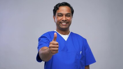 Wall Mural - healthcare, profession and medicine concept - happy smiling indian doctor or male nurse in blue uniform showing thumbs up over grey background