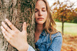 © iuricazac - Closeup portrait of a joyful young woman smiling, wearing a blue denim shirt embraces a tree, posing on nature background. Happy female enjoying the time in the park. Mental health concept.