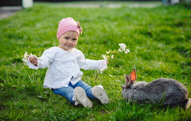 Little pretty girl playing with a rabbit in the garden. Cute baby in green grass on a sunny day. Portrait of one year old baby outdoors. Easter holidays concept. Easter bunny.