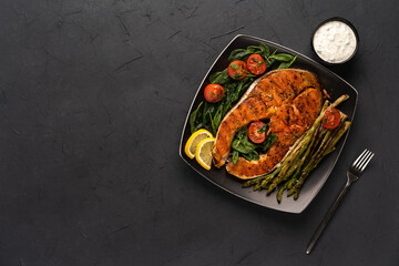 Wall Mural - grilled salmon steak, asparagus and fresh spinach with tomatoes on a black square plate on a dark background, space for text