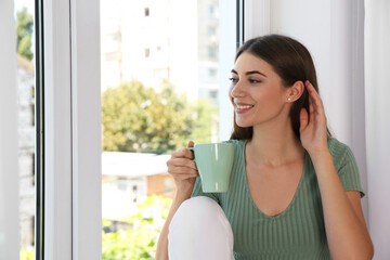 Poster - Beautiful young woman with cup of drink on window sill indoors