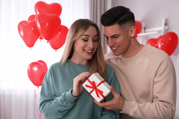 Poster - Man presenting gift to his girlfriend in room decorated with heart shaped balloons. Valentine's day celebration