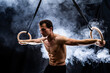 Muscular build man doing calisthenics on gymnastics rings indoor on black, smoked background. concept of motivation, desire and passion