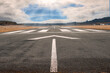 White arrow on a small asphalt air plane runway. Cloudy sky with small blue opening in the middle. Sun rays and flare. Small airport on the right. Aviation industry theme. Selective focus
