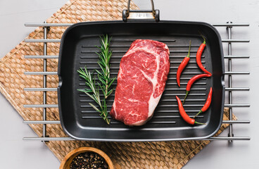 Wall Mural - Raw rib eye beef steak on black grilling pan with chili pepper and rosemary, top view.