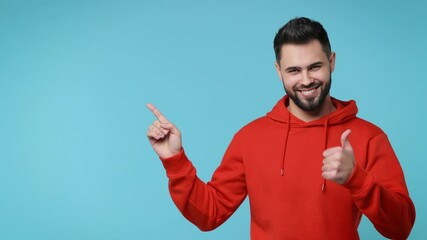 Wall Mural - Cheerful excited young man 20s years old in casual red streetwear hoodie isolated on blue background studio. People lifestyle concept. Pointing index finger aside up showing thumb up like gesture
