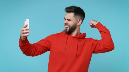 Wall Mural - Smiling funny young bearded man 20s in red hoodie isolated on blue background studio. People lifestyle concept. Doing selfie shot on mobile phone greeting with hand showing thumb up biceps muscles