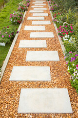 Selective focus shot of white stone path with yellow and brown gravel in colorful flower garden and green grass shows beautiful landscape of summer season. It is a gorgeous background of walkway