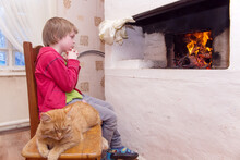 The Boy Sits Near The Stove And Looks At The Fire. The Cat Is Sleeping On A Chair. Selective Focus On The Boy.