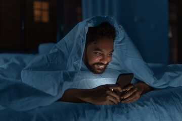 technology, internet, communication and people concept - young indian man with smartphone lying in bed under blanket at home at night