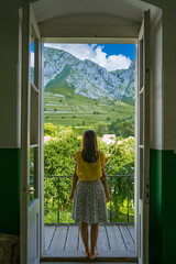 Inside a dark room with a woman looking into the distance through the wide-open doors that reveal the top of a mountain on a sunny summer day, a contrasting image that gives the idea of ​​hope.