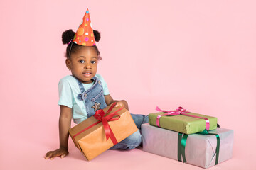 Poster - Indoor studio shot of cheerful pretty little African baby girl in casual clothes and red party hat, sitting on the floor with gift boxes, posing to camera. Isolated on pink background