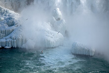 Freezing Mist Creating Ice Formations At The Bottom Of Niagara Falls, Ontario, Canada.  Seagulls Fly Through The Mist Above The River.