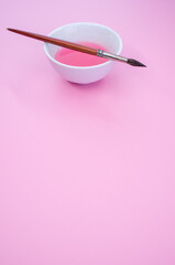 Wall Mural - Vertical shot of pink liquid watercolor paint on a bowl and paintbrush isolated on pink background