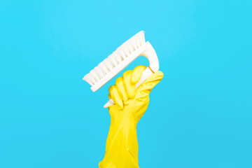 Hand in yellow glove holding up brush for cleaning surfaces, blue background. Equipment for washing and cleaning concept