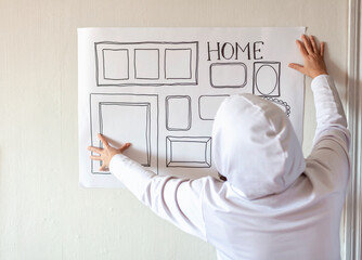 Woman evaluating and sizing up white poster with drawn set of different empty vertical and horizontal picture frames on white wall, planning family photo gallery project to capture moment, lifestyle