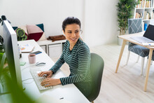 Smiling Young Businesswoman Looking Away While Sitting At Desk By Computer In Office