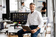 © peter scholl/Westend61 - Male entrepreneur with digital tablet sitting on desk with colleagues in background at open plan office