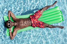 From above of carefree African American male with naked torso in shorts and sunglasses lying on inflatable mattress in swimming pool and enjoying sunny day during summer holiday