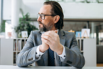 Portrait of a pensive senior male entrepreneur at the office. Serious middle aged businessman sits at the desk, looks away, thinking about solving work problems