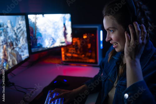 A girl with headphones plays a video game on a computer on large monitors. Gamer with mouse and keyboard. Online games with friends,win.Fun entertainment.Teenagers play adventure games.Neon lighting