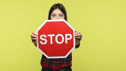 Wall Mural - Serious concentrated woman in checkered shirt holding in hands red stop sign, warning about danger, denies bad habit. Indoor studio shot isolated on yellow background