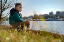 Cute Caucasian Boy Sitting On The Bank Of River In City Park Looking At Boats Passing By. Image With Selective Focus