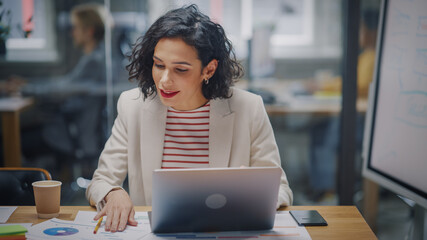 Aufkleber - Happy Latin Project Manager with Dark Wavy Hair Working on Laptop in Busy Creative Office. Beautiful Diverse Female Ad Manager Comparing Reports and Graphs on Paper and on Her Computer.