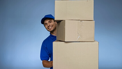 Wall Mural - cheerful delivery man holding carton boxes while smiling at camera isolated on blue