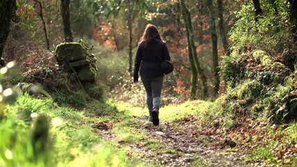 Wall Mural - Woman, with a black jacket with a hood, a backpack and grey pants, walking along a path, surrounded by vegetation, trees and stones, in a forest in Spain. Lifestyle concept, hiking and nature