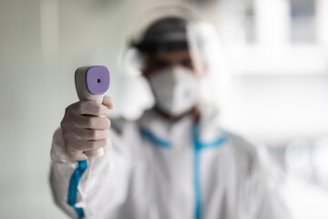 Portrait of man medic wearing biological protective uniform suit clothing, mask, gloves measuring body temperature with infrared forehead thermometer gun in hospital office