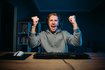 Portrait of a joyful gamer guy sitting at a table at home and rejoices in winning the game with his hands up and looks at the camera and shouts. Gaming concept