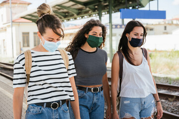 Three young beautiful female women at station to catch train for their vacation together during Coronavirus Covid-19 pandemic wearing protective face masks - Millennials have fun during the holidays