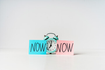 Alarm clock and a colored sticker with the inscription NOW on a grey background